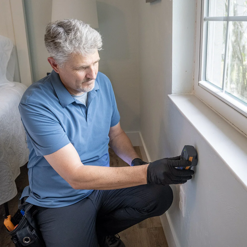 Kit Brucker kneeling by a window, holding a meter to the wall